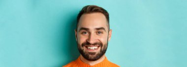Kameraya gülümseyen, turkuaz arka planda turuncu süveter giyen, sakallı, yakışıklı beyaz adamın vesikalık resmi..