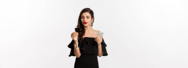 Moda ve alışveriş konsepti. Makyajlı ve siyah elbiseli çekici bir kadın online, kredi kartı ve cep telefonuyla ödeme yapıyor, beyaz arka plan.