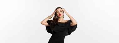 Siyah şık elbiseli, kendine güvenen ve şık bir kadın, kameraya küstah bakıyor, beyaz arka planda duruyor..