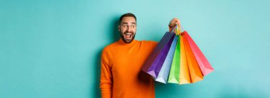 Mutlu genç adam alışverişe gidiyor, çantalarını tutuyor ve heyecanlı görünüyor, mavi arka planda turuncu süveter giyiyor..