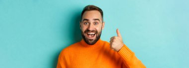 Neşeli erkek modelin yakın çekimi başparmağını kaldırıyor, gülümsüyor, açık mavi arka planda duruyor..