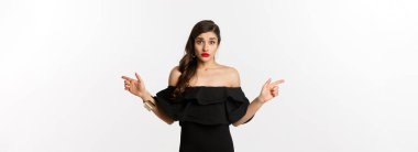 Moda ve güzellik. Belirsiz siyah elbiseli kadın parmaklarını yana doğru gösteriyor, iki seçenek gösteriyor ve kafası karışık, beyaz arka plan.