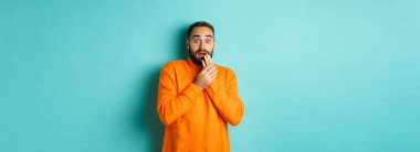 Korkmuş ve şok olmuş bir adam, turkuaz arka planda turuncu kazak giymiş kameraya bakıyor..