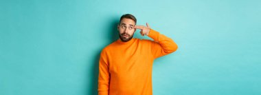 Adam kendini kafasından vuruyor, parmak tabancasını şakağa yaklaştırıyor ve gözleri yuvarlanıyor, açık mavi arka plana karşı koymaktan rahatsız oluyor..