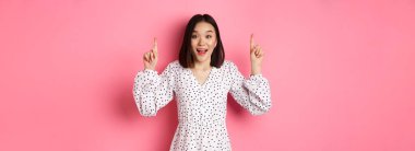 Şaşırmış esmer Asyalı kız parmak kaldırıyor, reklam yapıyor, bahar tanıtım teklifini sunuyor, pembe arka planda duruyor..