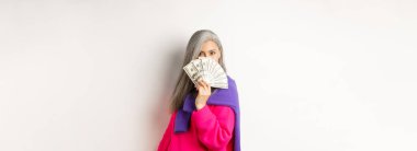 Alışveriş konsepti. Zengin ve modaya uygun Koreli son sınıf öğrencisi bir kadın paranın arkasına saklanıyor, kameraya bakıyor, beyaz arka planda duruyor..