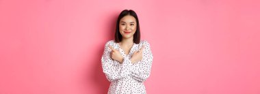 Romantik elbiseli güzel Asyalı kız, iki farklı alışveriş çeşidini gösteriyor, kameraya gülümsüyor, pembe arka planda duruyor..