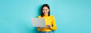 Genç Asyalı kadın dizüstü bilgisayarla çalışıyor ve gülümsüyor, mavi arka planda bilgisayarın başında duruyor..