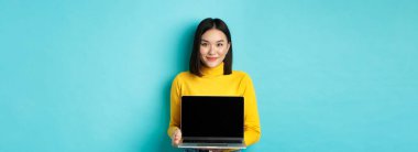 Güzel ve stil sahibi Asyalı kadın ürününü ekranda gösteriyor. Boş dizüstü bilgisayarını gösteriyor ve gülümsüyor. Mavi arka planda duruyor..