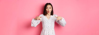Etkilenmiş Asyalı kadın parmakları aşağıda, şuraya bak, indirimlere ve satışlara bak, pembe arka planda şaşkın şaşkın duruyor..
