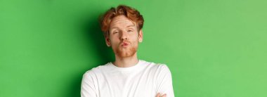 Duygular ve moda konsepti. Aptal kızıl saçlı, sakallı, dudaklarını büzüştüren ve öpüşmek için eğilen yeşil arka planda duran adam..