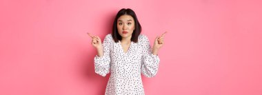 Kafası karışmış Asyalı kadın tereddüt ediyor, omuz silkiyor ve iki tarafı işaret ediyor, kararsız bakıyor ve seçiminde yardıma ihtiyacı var, pembe arka planda duruyor..