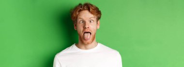 Duygular ve moda konsepti. Komik, kızıl saçlı, aptal suratlar gösteren, dil uzatan ve gözlerini kısan, yeşil arka planda duran adamın yakın çekimi..