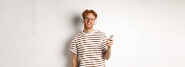 Teknoloji konsepti. Kızıl saçlı ve sakallı genç adam kulaklıkla müzik dinliyor ve akıllı telefon kullanıyor, kameraya gülümsüyor, beyaz arka plan.