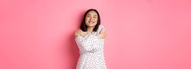 Güzel Asyalı bir kadın romantik hissediyor, kendine sarılıyor, gözlerini kapatıyor ve rüya gibi yüzüyle gülümsüyor, pembe arka planda mutlu bir şekilde duruyor..