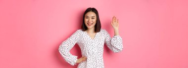 Güzel Asyalı kadın merhaba diyor, selamlaşmak için el sallıyor ve merhaba diyor, kameraya gülümsüyor, pembe arka planda duruyor..