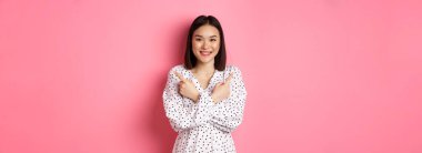 Romantik elbiseli güzel Asyalı kız, iki farklı alışveriş çeşidini gösteriyor, kameraya gülümsüyor, pembe arka planda duruyor..