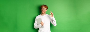 Gözlüklü yakışıklı genç erkek. Tamam işareti, dizüstü bilgisayarı var. Yeşil arka planda duruyor..