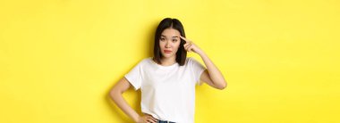 Sinirli genç Asyalı bir kadın aptal birini azarlıyor, parmağıyla kafasını işaret ediyor, deli misin diye soruyor, sarı arka planda dikiliyor..