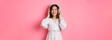 Romantik Asyalı kadın mutlu gülümsüyor, kulaklıkla müzik dinliyor ve pembe arka planda duran kameraya bakıyor..