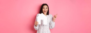 Gayrimenkul. Heyecanlı Asyalı kadın kağıt ev modelini gösteriyor, fotokopi alanına bakıyor ve sola bakıyor, pembe arka planda duruyor..