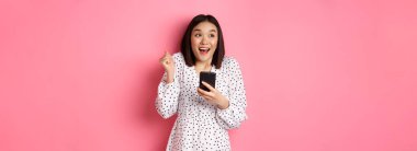 İnternetten alışveriş ve güzellik konsepti. Heyecanlı Asyalı kadın internette kazanıyor, elinde akıllı telefon, gülümsüyor, mutlu ve mutlu, pembe arka planda duruyor..