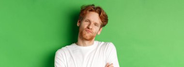 Duygular ve moda konsepti. Kızıl saçlı, sakallı, büzüşmüş dudakları olan, kameraya bakan, yeşil arka planda duran şüpheci genç adamın yakın çekimi..