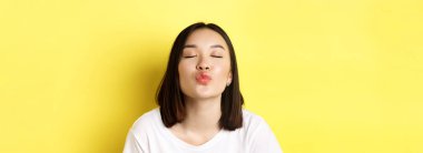 Sevgililer Günü konsepti. Tatlı Asyalı dudaklarını büzen dudaklarını ve öpmek için gözlerini kapatan, sarı arka planda romantik duran.