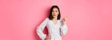 Güzel Asyalı kız sol üst köşeyi işaret ediyor. Kafası karışmış ve şüpheli görünüyor. Pembe arka planda duruyor..