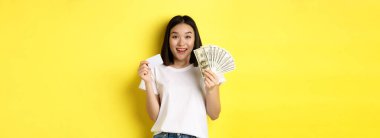 Kısa siyah saçlı, beyaz tişörtlü, parayı dolar ve kredi kartlarıyla gösteren, sarı arka planda duran güzel Asyalı kadın..