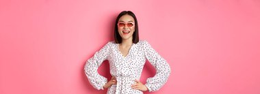 Turizm ve yaşam tarzı konsepti. Güzel Koreli kadın tatilde, güneş gözlüğü ve elbisesi takıyor, mutlu bir gülümsemeyle sola bakıyor, pembe arka planda duruyor..