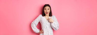 Güzel esmer Asyalı kadın kamerayı işaret ediyor. Seçilmiş kişi olarak şaşırmış görünüyor. Pembe arka plana karşı giyinmiş..