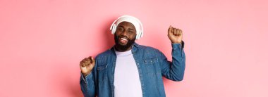 Mutlu siyah adam dans ediyor ve kulaklıkla müzik dinliyor. Neşeli gülümsüyor, sokak tarzı kıyafetlerle pembe arka planda duruyor..
