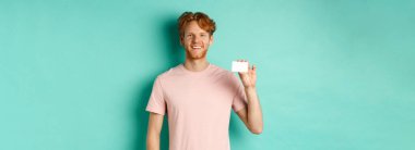 Yakışıklı genç adam gülümsüyor ve plastik kredi kartı gösteriyor. Tişörtünün içinde turkuaz arka planda duruyor..