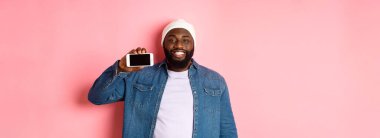 Çevrimiçi alışveriş ve teknoloji kavramı. Yakışıklı siyahi adam akıllı telefon ekranını yatay olarak gösteriyor ve gülümsüyor tatmin olmuş, uygulama öneriyor, pembe arka plan.