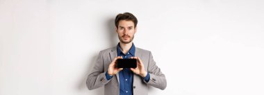 E-ticaret ve online alışveriş konsepti. Takım elbiseli ciddi genç adam boş akıllı telefon ekranını yatay gösteriyor, beyaz arka planda duruyor..