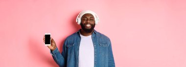 Mutlu hipster adam kulaklıkla müzik dinliyor ve mobil ekran gösteriyor, gülümsüyor, pembe arka planda duruyor..