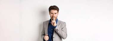 Kızgın işadamı tabu işareti gösteriyor, sessiz olmasını söylüyor, sessizliğe ihtiyacı var, parmaklarını dudaklarına dayayıp kameraya bakıyor, beyaz arka plan.