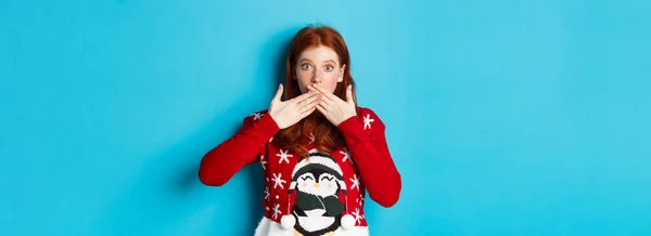 Winter holidays and Christmas Eve concept. Surprised redhead girl in cute sweater, gasping and covering mouth with hands, staring at camera, standing over blue background.