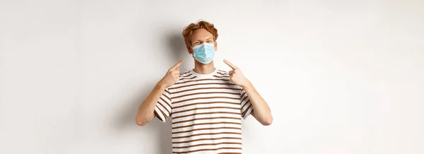 Covid, virus and social distancing concept. Smiling redhead guy pointing fingers at face mask, standing over white background.