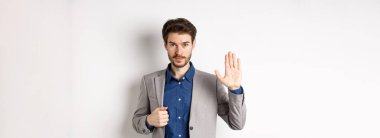 Resmi takım elbiseli bir iş adamı merhaba demek için elini kaldırıyor. Temas kurmadan selamlıyor, gülümsüyor, beyaz arka planda duruyor..