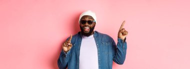 Mutlu sakallı Afro-Amerikalı adam anons yapıyor, sol üst köşeyi işaret ediyor ve gülümsüyor, bereyle ve pembe arka planda güneş gözlüğü takıyor..