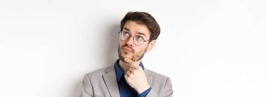 Sakallı ve gözlüklü düşünceli bir işadamı, karar veriyor, yukarı bakıyor ve düşünüyor, düşünceli yüzüyle takım elbise içinde duruyor, beyaz bir geçmişi var..