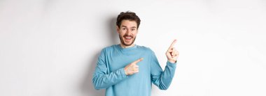 Mavi kazaklı mutlu bir beyaz adam, sol üst köşeyi işaret ediyor, beyaz arka planda bağlantı ya da logo gösteriyor, kameraya gülümsüyor..