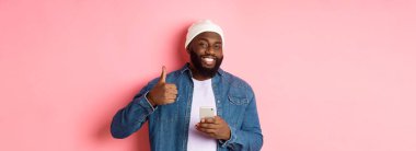 Teknoloji ve online alışveriş konsepti. Mutlu Afro-Amerikalı adam cep telefonu kullanırken onay veriyor, pembe arka planda tatmin olmuş duruyor..