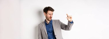 İş adamı küçük bir şey görmeye çalışıyor, parmaklarıyla küçük bir şey gösteriyor, beyaz arka planda takım elbise içinde duruyor..