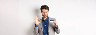 Plastik kredi kartı ve tamam işareti gösteren mutlu bir erkek banka müşterisi. Gülümsüyor, beyaz arka planda duruyor..