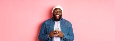 Teknoloji ve online alışveriş konsepti. Şaşkın genç siyahi adam cep telefonu kullanıyor, kameraya bakıyor. Mesajı okuduktan sonra şaşkın ve mutlu. Pembe arka plan..