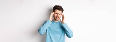 Genç adam baş ağrısı çekiyor ya da akşamdan kalma, baş şakaklarına dokunuyor ve kaşlarını çatıyor, migrenden midesi bulanıyor, beyaz arka planda duruyor. .