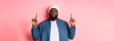 Ciddi ve canı sıkkın sakallı siyahi adam, kameraya bakıyor ve işaret ediyor, reklamını yapıyor, pembe arka planda duruyor..